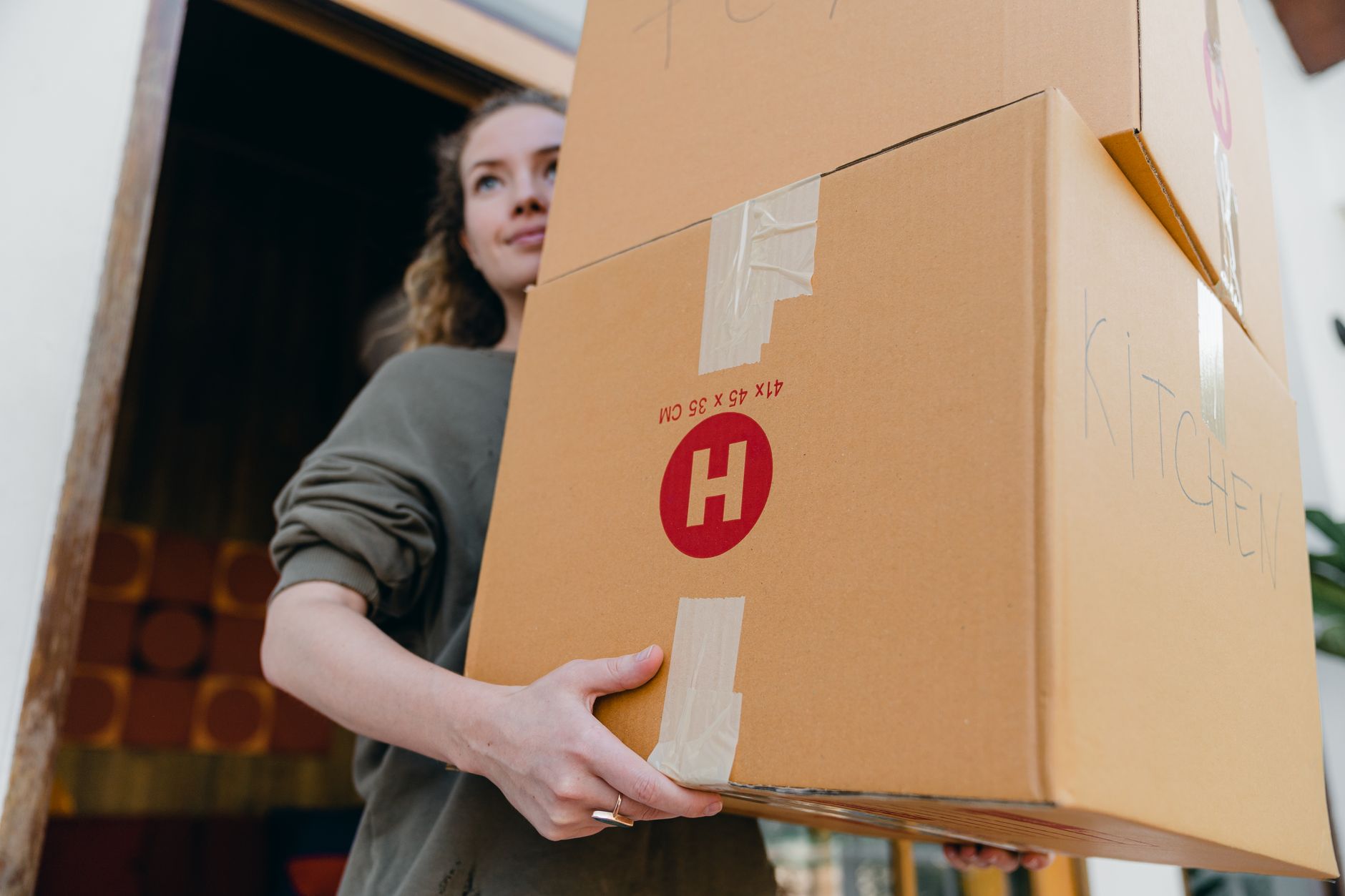 From below of young slim woman carrying stack of heavy carton boxes with kitchen stuff and looking away while moving out of o